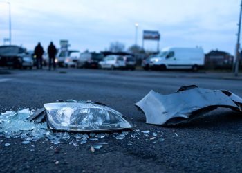 Shattered headlight lens and broken bumper metal in sharp focus on dark pre-dawn pavement, with damaged and emergency vehicles blurred behind at a Commerce intersection.