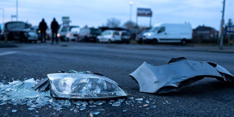 Shattered headlight lens and broken bumper metal in sharp focus on dark pre-dawn pavement, with damaged and emergency vehicles blurred behind at a Commerce intersection.
