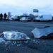 Shattered headlight lens and broken bumper metal in sharp focus on dark pre-dawn pavement, with damaged and emergency vehicles blurred behind at a Commerce intersection.