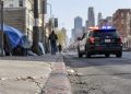 Skid Row street in downtown Los Angeles with brick buildings and chain-link fencing, street curb in sharp focus, patrol SUV further down, softly blurred.
