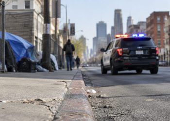 Skid Row street in downtown Los Angeles with brick buildings and chain-link fencing, street curb in sharp focus, patrol SUV further down, softly blurred.