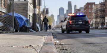 Skid Row street in downtown Los Angeles with brick buildings and chain-link fencing, street curb in sharp focus, patrol SUV further down, softly blurred.
