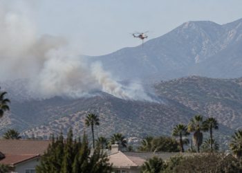 Smoke rising over dry brush-covered foothills above a residential neighborhood in Azusa in afternoon daylight, with a firefighting helicopter in the distance.