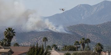 Smoke rising over dry brush-covered foothills above a residential neighborhood in Azusa in afternoon daylight, with a firefighting helicopter in the distance.