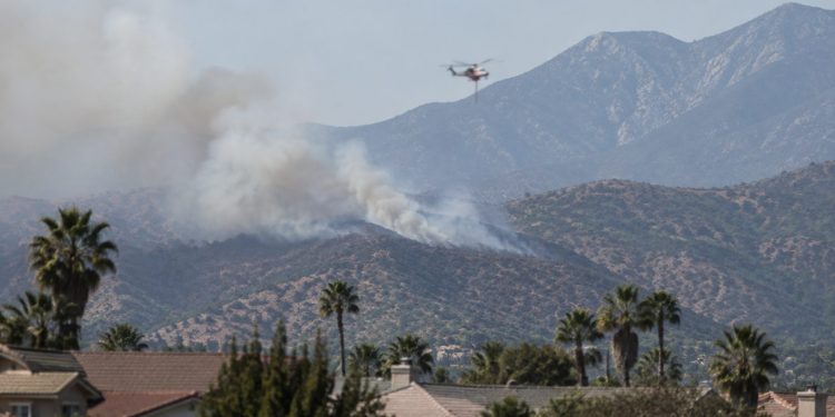 Smoke rising over dry brush-covered foothills above a residential neighborhood in Azusa in afternoon daylight, with a firefighting helicopter in the distance.