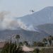 Smoke rising over dry brush-covered foothills above a residential neighborhood in Azusa in afternoon daylight, with a firefighting helicopter in the distance.