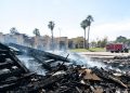 Smoldering remains of a single-story strip mall in Inglewood under midday sun, with charred debris in the foreground and a fire engine blurred behind.