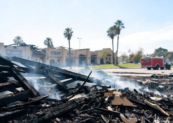 Smoldering remains of a single-story strip mall in Inglewood under midday sun, with charred debris in the foreground and a fire engine blurred behind.