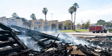 Smoldering remains of a single-story strip mall in Inglewood under midday sun, with charred debris in the foreground and a fire engine blurred behind.