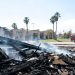Smoldering remains of a single-story strip mall in Inglewood under midday sun, with charred debris in the foreground and a fire engine blurred behind.