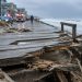 Splintered boardwalk planks in sharp focus on Seaside Walk, with oceanfront homes and the churning sea blurred in daylight on the Long Beach Peninsula.