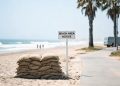 Stacked sandbags on a Southern California beach in hot, hazy daylight, with 3-to-5-foot surf breaking in the blurred background.