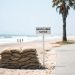 Stacked sandbags on a Southern California beach in hot, hazy daylight, with 3-to-5-foot surf breaking in the blurred background.