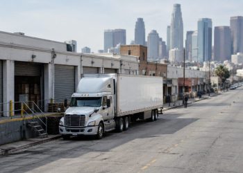 Tractor-trailer parked beside low-rise wholesale buildings with roll-up metal doors on a downtown Los Angeles street in neutral daylight, towers blurred behind.
