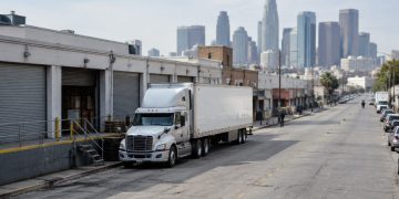Tractor-trailer parked beside low-rise wholesale buildings with roll-up metal doors on a downtown Los Angeles street in neutral daylight, towers blurred behind.