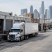 Tractor-trailer parked beside low-rise wholesale buildings with roll-up metal doors on a downtown Los Angeles street in neutral daylight, towers blurred behind.