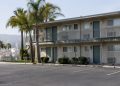 Two-story motel exterior in Los Angeles County with a second-floor walkway and small bathroom windows under neutral daylight, palm trees beside an empty lot.