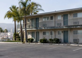Two-story motel exterior in Los Angeles County with a second-floor walkway and small bathroom windows under neutral daylight, palm trees beside an empty lot.