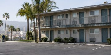 Two-story motel exterior in Los Angeles County with a second-floor walkway and small bathroom windows under neutral daylight, palm trees beside an empty lot.