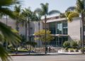 West Hollywood City Hall seen from across Santa Monica Boulevard in neutral daylight, with palm trees in the foreground, illustrating the city council race.