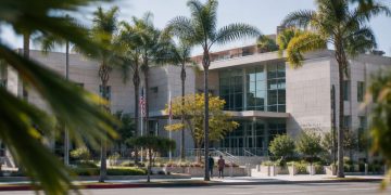 West Hollywood City Hall seen from across Santa Monica Boulevard in neutral daylight, with palm trees in the foreground, illustrating the city council race.
