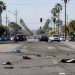 Wide view of a South Los Angeles intersection with vehicle debris scattered across the asphalt, low-rise storefronts and palm trees in morning daylight.