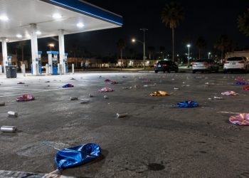 Wide view of a gas station parking lot at night in Bellflower, with small silver canisters and balloons scattered across the asphalt under canopy lights.