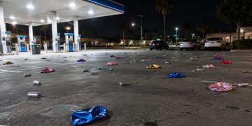 Wide view of a gas station parking lot at night in Bellflower, with small silver canisters and balloons scattered across the asphalt under canopy lights.