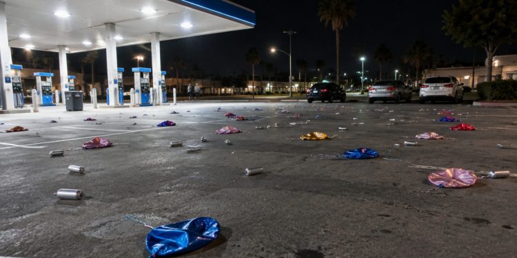 Wide view of a gas station parking lot at night in Bellflower, with small silver canisters and balloons scattered across the asphalt under canopy lights.