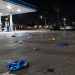 Wide view of a gas station parking lot at night in Bellflower, with small silver canisters and balloons scattered across the asphalt under canopy lights.