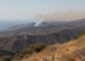 Wide view of dry chaparral ridgelines in the Santa Monica Mountains near Malibu with a thin plume of smoke rising over the hills in afternoon daylight.