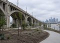 Wide view of newly graded park grounds with young trees and paved paths beneath the arched concrete spans of a large Los Angeles viaduct in daylight.
