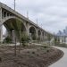 Wide view of newly graded park grounds with young trees and paved paths beneath the arched concrete spans of a large Los Angeles viaduct in daylight.