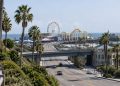 Wide view of the Santa Monica Pier entrance in neutral daylight, with palm trees along Ocean Avenue and the Pacific Park Ferris wheel softly blurred beyond.