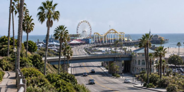 Wide view of the Santa Monica Pier entrance in neutral daylight, with palm trees along Ocean Avenue and the Pacific Park Ferris wheel softly blurred beyond.