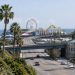 Wide view of the Santa Monica Pier entrance in neutral daylight, with palm trees along Ocean Avenue and the Pacific Park Ferris wheel softly blurred beyond.