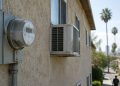 Window air-conditioning units and a utility meter on the side of an older Los Angeles apartment building in afternoon daylight, illustrating high utility bills.