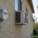 Window air-conditioning units and a utility meter on the side of an older Los Angeles apartment building in afternoon daylight, illustrating high utility bills.